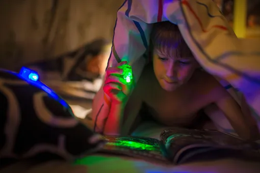 мальчик с фонариком читает книгу под одеялом