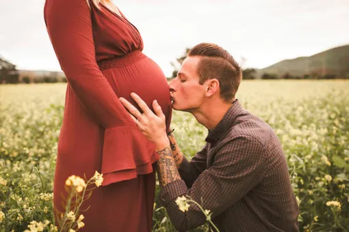 на лугу мужчина целует живот беременной женщины в красном платье