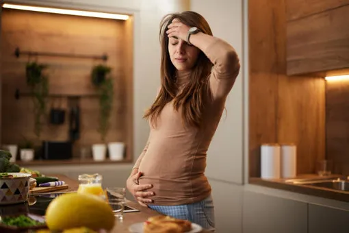 беременная женщина стоит на кухне, одной рукой держится за живот, другой за голову