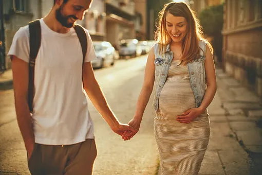 беременная женщина идет рядом с мужем, придерживает живот, они держатся за руки