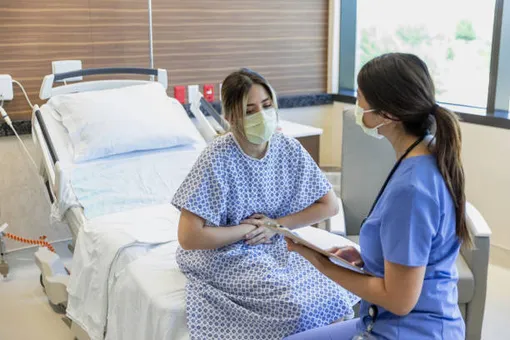 врач в маске разговаривает с женщиной в больничной рубашке и маске, сидящей на кровати и держащейся за животмаске