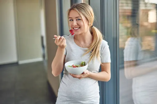беременная женщина стоит и ест овощи из миски