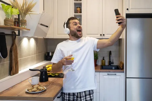 Мужчина с бокалом сока в наушниках на кухне смотрит в экран телефона и смеется, радуется пожеланию доброго утра