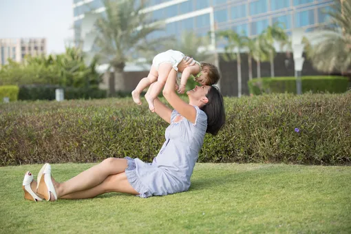 мама сидит на газоне и подняла на руках ребенка