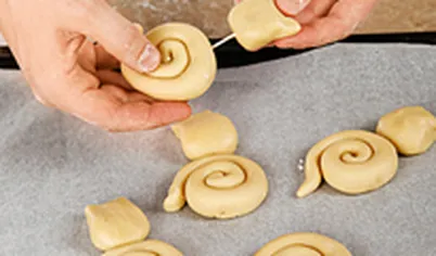 Из теста скатайте колбаску, разрежьте на 12 одинаковых частей размером с грецкий орех и 12 частей большего размера. Из больших кусочков скатайте жгутики и закрутите спиралькой. Из мелких кусочков слепите «головы» котят с ушками. С помощью зубочистки закрепите детали и выпекайте в разогретой до 180 градусов духовке на выстеленном пекарской бумагой противне примерно 10 минут.