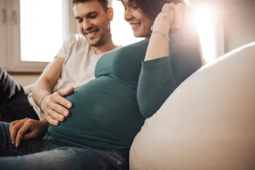 беременная женщина сидит на диване, мужчина сидит рядом, положив ладонь ей на живот