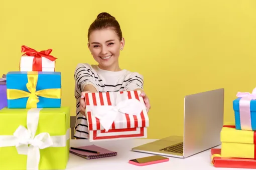 Девушка перед ноутбуком улыбается и держит коробку с подарком и рядом стоит много подарочных коробок Поздравления с 8 марта коллегам