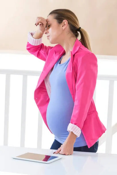 беременная женщина в розовом пиджаке держится за голову, на столе лежит планшет