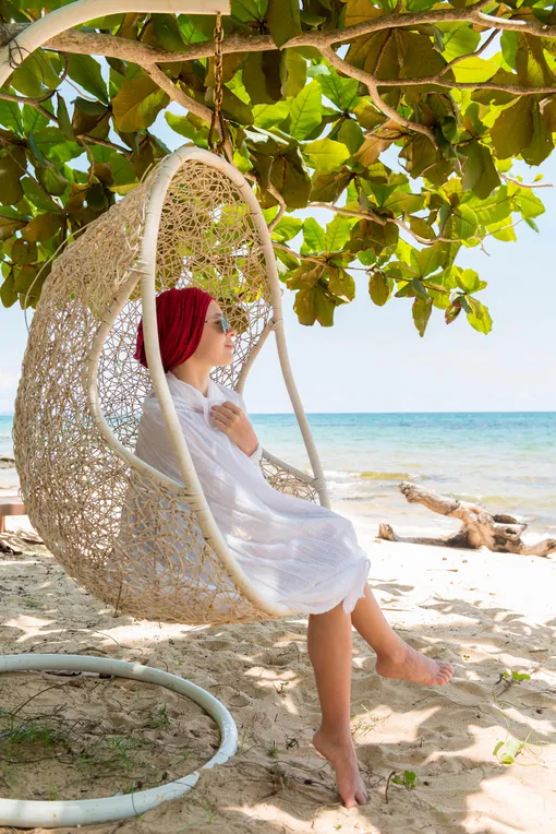 Девушка в плетеном кресле-яйце, подвешенном на ветку дерева на пляже
