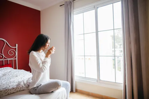 Девушка сидит на кровати, пьет кофе и смотрит в окно (ей пожелали доброго утра)