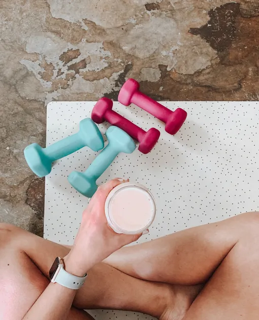 Розовые и голубые гантели и рука с белым спортивным напитком в стакане