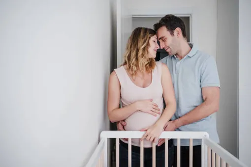 беременная женщина и ее мужчина стоят у детской кровати, мужчина ее обнимает за живот