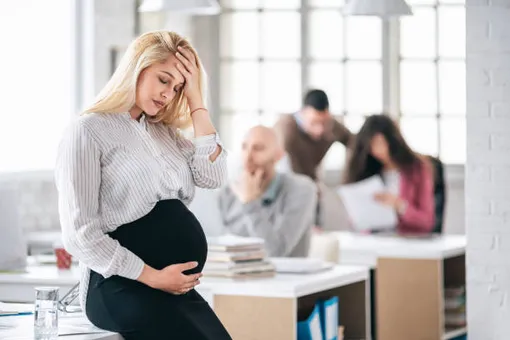 беременная женщина в офисе стоит и держится одной рукой за голову, другой за живот