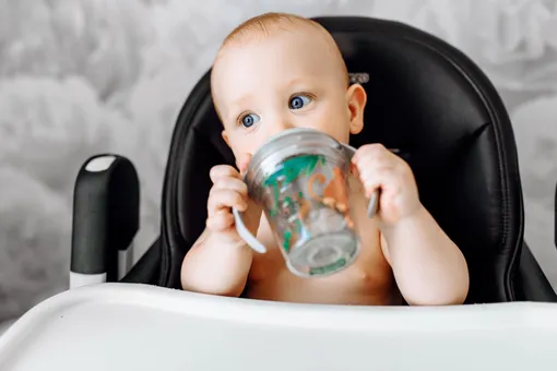 Малыш в стульчике для кормления с чашкой-непроливайкой в руках