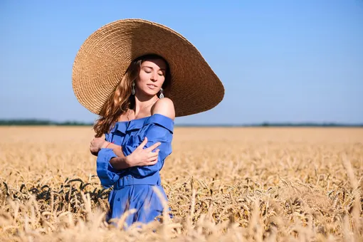 Девушка в поле пшеницы в синем платье и широкополой шляпе сама себя обнимает