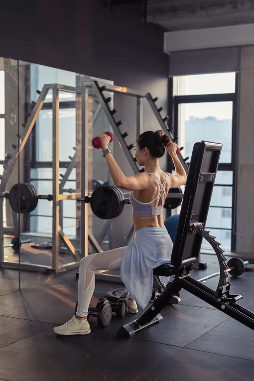 Девушка в белых лосинах и спортивной юбке делает упражнения с гантелями в тренажерном зале