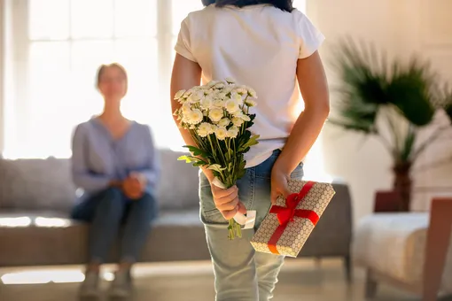 Дочка держит за спиной цветы и подарок для мамы сидящей на диване Поздравления с 8 марта маме