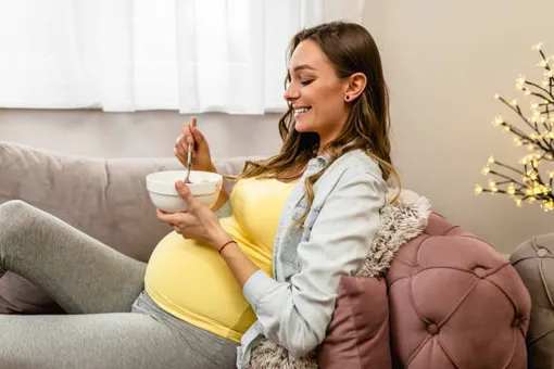 беременная женщина сидит на диване и ест что-то из миски