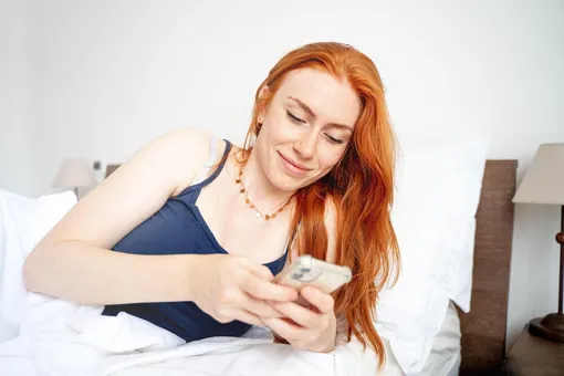 Рыжая девушка в кровати читает на телефоне пожелания с добрым утром