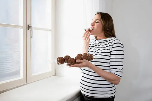 беременная женщина стоит у окна и ест пончики