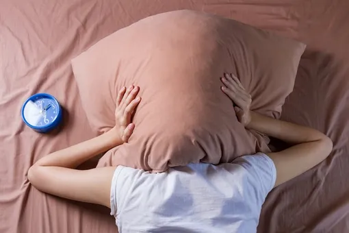 Девушка в белой футболке накрылась подушкой в попытках наладить сон, который помогает худеть без диет и спорта