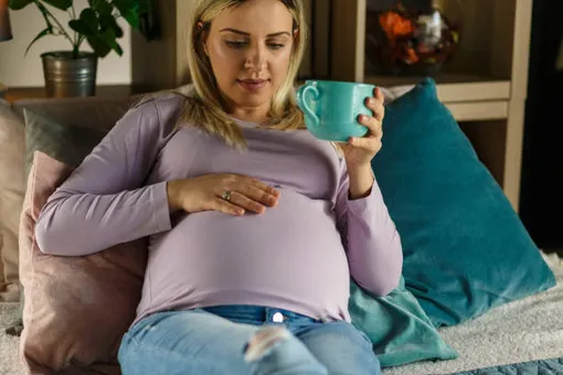 беременная женщина сидит в кресле с кружкой в руках и смотрит на живот, положив на него рукуу