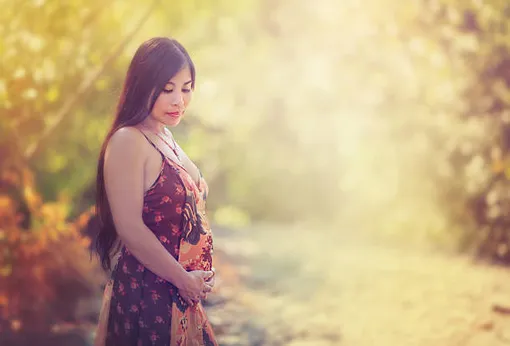 беременная женщина с длинными волосами стоит на фоне природы и придерживает живот